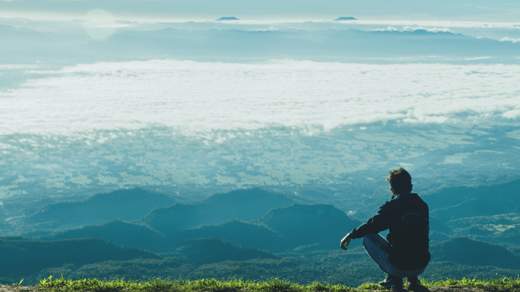 A person squatting on a mountain ledge overlooking a vast valley covered by clouds, symbolising reconnection to natural movement and body awareness.